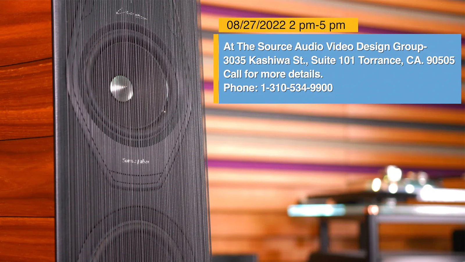 High-end speaker at The Source AV showroom with event info overlay, wood panel background
